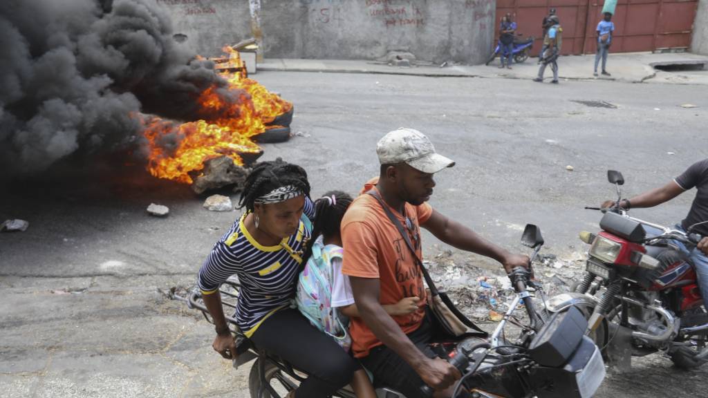ARCHIV - Ein Motorradtaxifahrer transportiert in Port-au-Prince, Haiti, Kunden vorbei an Reifen, die von Demonstranten in Brand gesteckt wurden, die den Rücktritt des Übergangspräsidentenrates forderten. Foto: Odelyn Joseph/AP/dpa