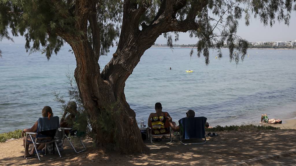 Besucher sitzen im Schatten unter einem Pinienbaum am Strand des Vororts Kavouri südwestlich von Athen. Foto: Yorgos Karahalis/AP/dpa
