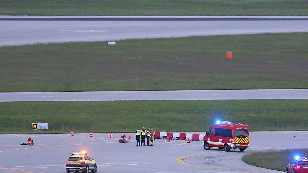 Einsatzkräfte von Polizei und Feuerwehr stehen um Klimaaktivisten, die sich am Münchner Flughafen auf eine Start- beziehungsweise Landebahn festgeklebt haben. Foto: Karl-Josef Hildenbrand/dpa