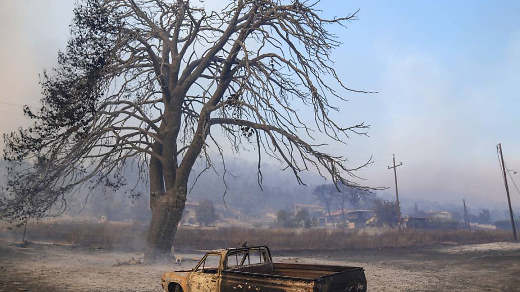 Ein beschädigtes Auto steht vor einem verbrannten Baum in der Nähe von Loutraki. Nach einer längeren Trockenperiode sind im Großraum der griechischen Hauptstadt Athen mehrere große Wald- und Buschbrände ausgebrochen. Foto: Petros Giannakouris/AP/dpa