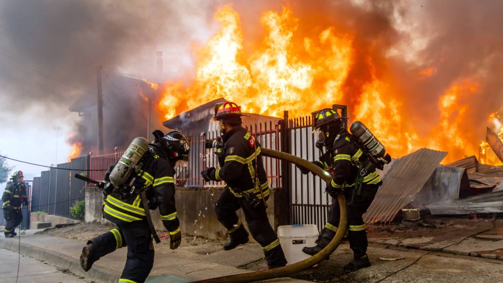 Feuerwehrleute bekämpfen ein Feuer in einem Haus, während Waldbrände lodern. Foto: Javier Torres/AP/dpa