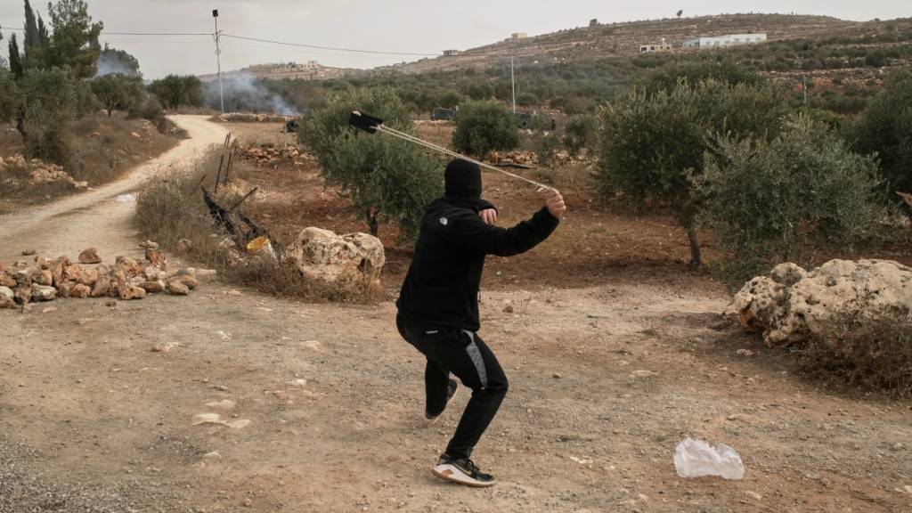 Ein Palästinenser schleudert mit einer Schleuder einen Stein, während Tränengas zwischen den Olivenbäumen weht - bei Zusammenstößen nach einem Angriff von Siedlern auf Palästinenser, die im Dorf Abu Falah im Westjordanland Oliven pflückten. Foto: Leo Correa/AP/dpa