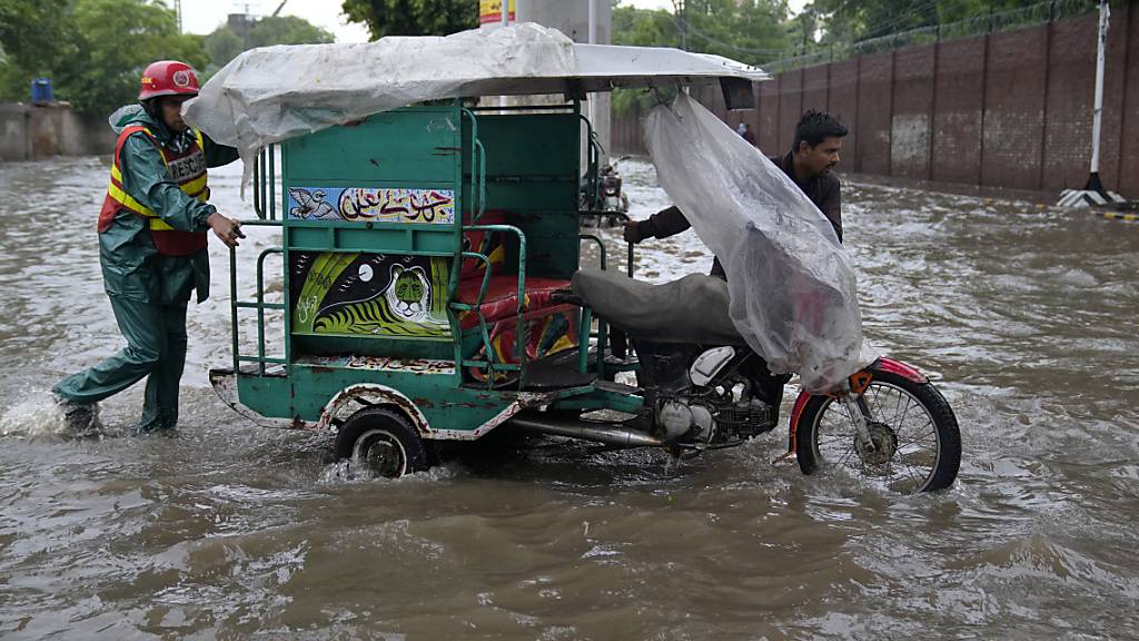 Ein Mitglied eines Rettungsteams hilft einem Mann seine Motorrad-Rikscha über eine überschwemmte Straße zu schieben. Schwere Monsunregenfälle haben in Pakistan nach offiziellen Angaben bereits mehrere Todesopfer gefordert. Foto: K.M. Chaudary/AP/dpa