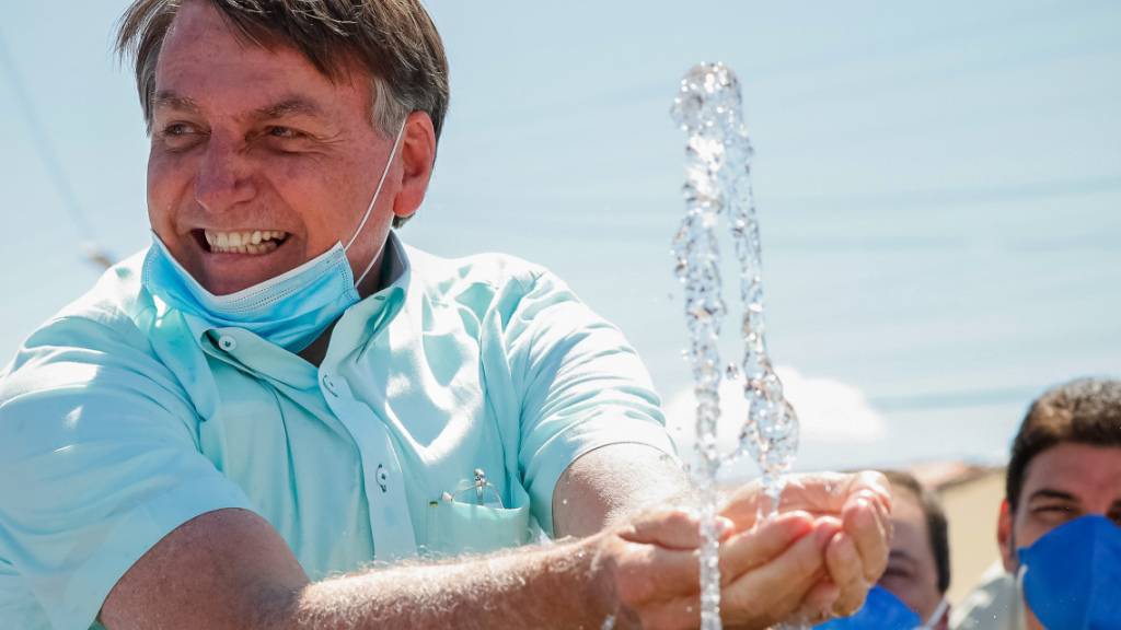 HANDOUT - Auf diesem vom brasilianischen Präsidentenamt zur Verfügung gestellten Bild lächelt Jair Bolsonaro, Präsident von Brasilien, mit untergezogenem Mundschutz, während er an einem Brunnen zur lokalen Wasserversorgung mit Wasser spielt. Foto: Alan Santos/Palacio Planalto/dpa