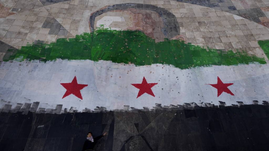 ARCHIV - Ein syrischer Junge steht unter einem riesigen Porträt des verstorbenen syrischen Präsidenten Al-Assad, übermalt mit den Farben der sogenannten «revolutionären» Flagge, in Damaskus. Foto: Hussein Malla/AP/dpa