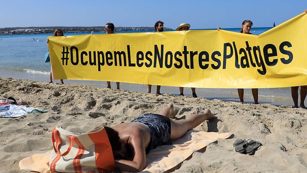 Leute halten an einem Strand auf Mallorca ein Banner «#Sie besetzen unsere Strände» aus Protest gegen den Massentourismus auf der Balearen-Insel. Foto: Clara Margais/dpa