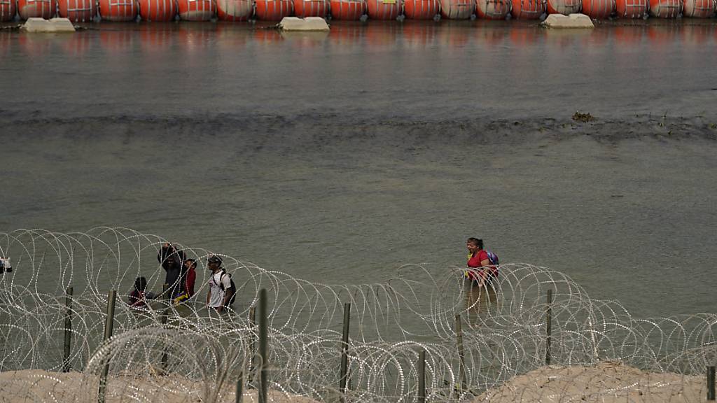 Migranten aus Kolumbien gehen entlang des Concertina-Stacheldrahts und einer schwimmenden Bojenbarriere, nachdem sie den Rio Grande von Mexiko in die USA überquert haben. Die schwimmende Grenze soll Menschen daran hindern, in die USA zu kommen. Foto: Eric Gay/AP/dpa