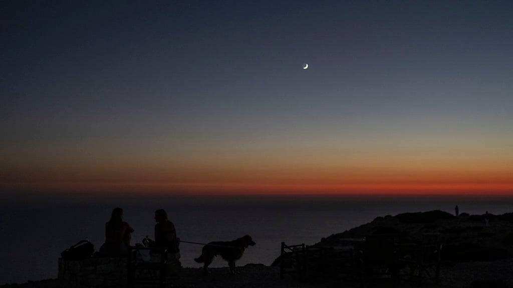 ARCHIV - Zwei Frauen mit einem Hund schauen auf das Libysche Meer, während der Mond über der griechischen Insel Gavdos am Himmel steht. Foto: Petros Giannakouris/AP/dpa