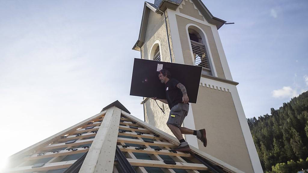 Arbeiter montieren Solarpanels auf dem Dach einer historischen Kirche im bündnerischen Trin. Das kantonale Aktionsprogramm Green Deal, das Klimaschutz und Gebäudesanierungen fördert, wurde vom Grossen Rat mit einem millionenschweren Zusatzkredit vor dem Stocken bewahrt.