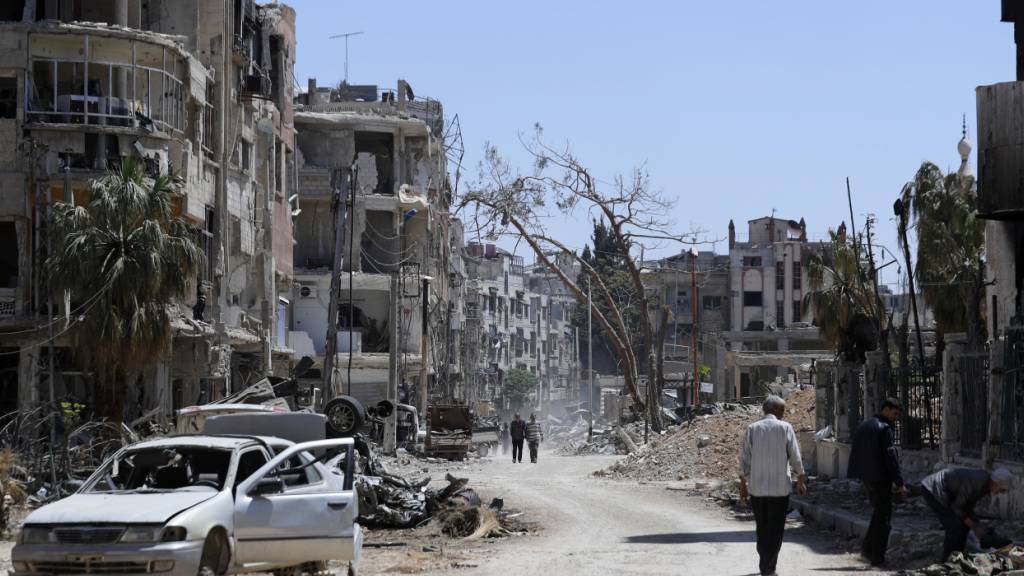 ARCHIV - 16.04.2018, Syrien, Duma: Menschen gehen an zerstörten Häusern und Autos im Vorort der syrischen Hauptstadt Damaskus vorbei. Foto: Hassan Ammar/AP/dpa