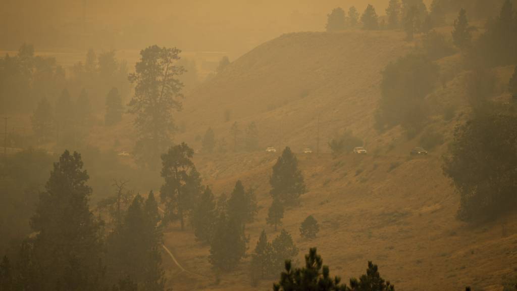Rauch von Waldbränden füllt die Luft, während Autofahrer auf einer Straße am Berghang in Kelowna, British Columbia, fahren. Foto: Darryl Dyck/The Canadian Press via AP/dpa - ACHTUNG: Nur zur redaktionellen Verwendung und nur mit vollständiger Nennung des vorstehenden Credits