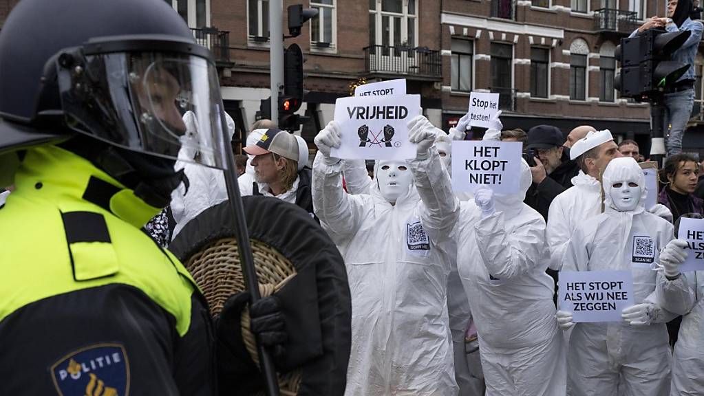 Demonstranten halten Plakate hoch, während ein Bereitschaftspolizist im Vordergrund steht. In Amsterdam hat die Polizei eine verbotene Demonstration gegen die Corona-Politik der Regierung aufgelöst..Am Sonntag hatten nach Angaben der Nachrichtenagentur ANP rund 2000 Menschen demonstriert. Foto: Peter Dejong/AP/dpa