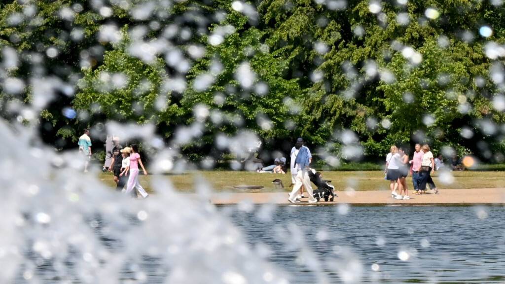 Menschen laufen bei sommerlichen Temperaturen durch die Kensington Gardens, während ein kleiner Springbrunnen Wasser versprüht. Foto: Anna Ross/dpa