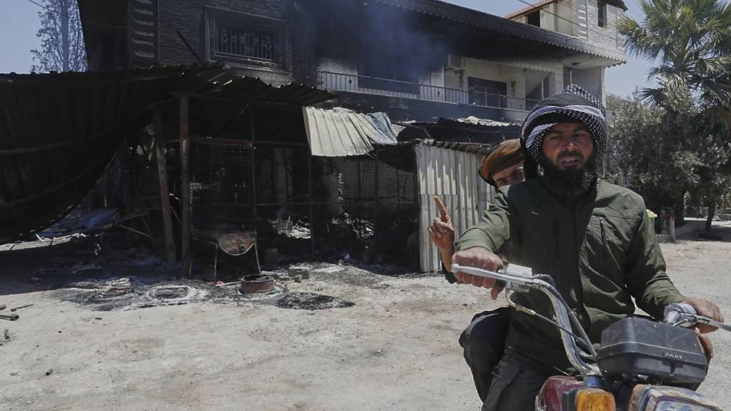 Zwei Syrer fahren auf einem Motorrad an einem qualmenden Haus in der syrischen Provinz Suwaida vorbei. Foto: Omar Sanadiki/AP/dpa