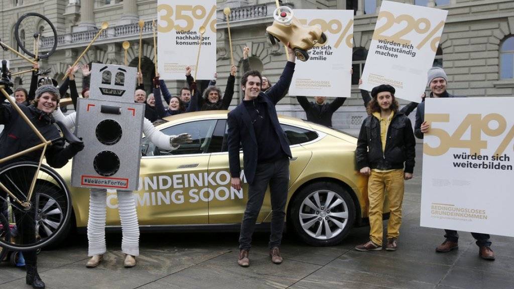 Die Initianten lancieren die Abstimmungskampagne für ein bedingungsloses Grundeinkommen auf dem Bundesplatz in Bern - vor einem goldfarbenen Tesla Auto und mit Plakaten zu den Umfrageergebnissen.