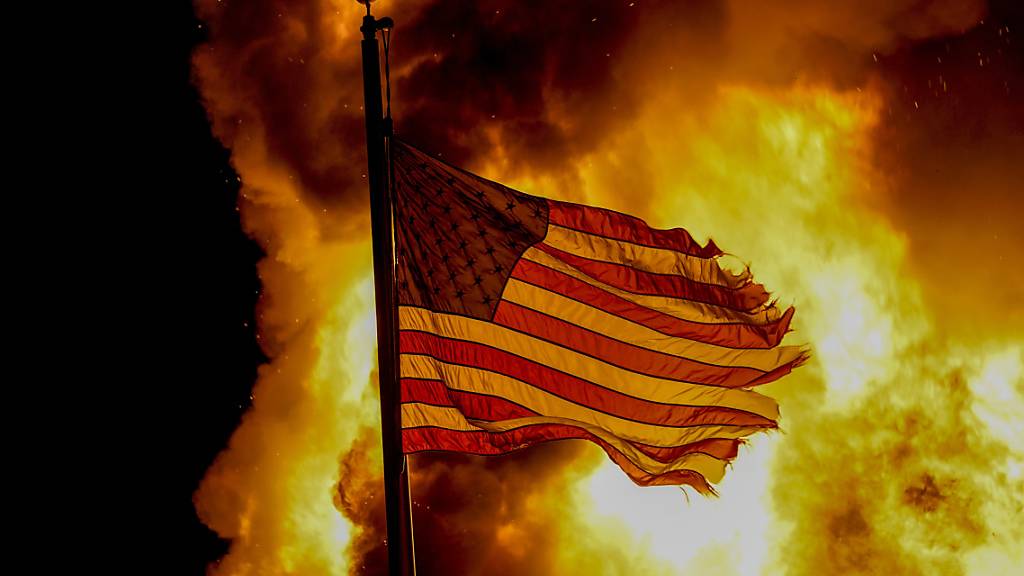 Eine ausgefranste US-Flagge weht vor den Flammen eines Feuers bei einem Protest nach den Schüssen auf Jacob Blake. Foto: Morry Gash/AP/dpa