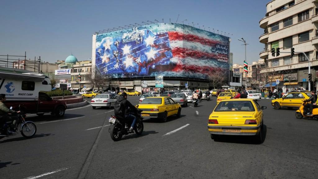Fahrzeuge fahren an einer Werbetafel vorbei, auf der ein US-Flugzeugträger mit beschädigten Kampfjets an Deck abgebildet ist. Auf dem Enqelab-e-Eslami-Platz (Platz der Islamischen Revolution) steht in Farsi und Englisch: «Wer Wind sät, wird Sturm ernten». Foto: Vahid Salemi/AP/dpa